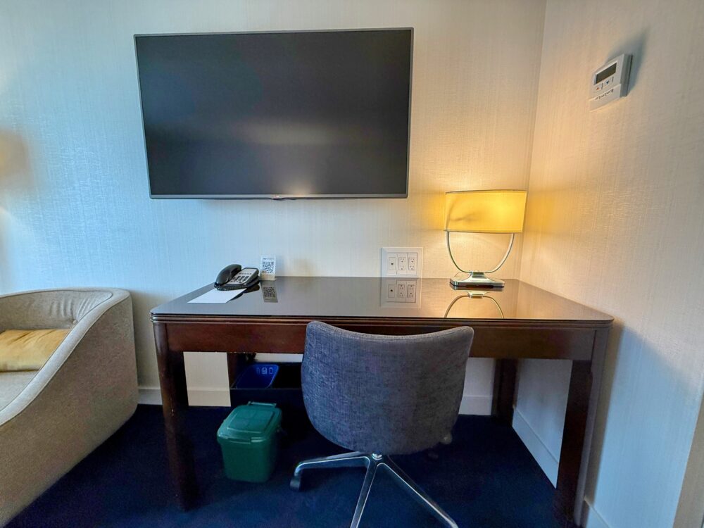 機能的なワークデスクとグレーのオフィスチェア。壁には大型の液晶テレビ、デスク上には照明と電話が設置され、ビジネス利用にも適している。