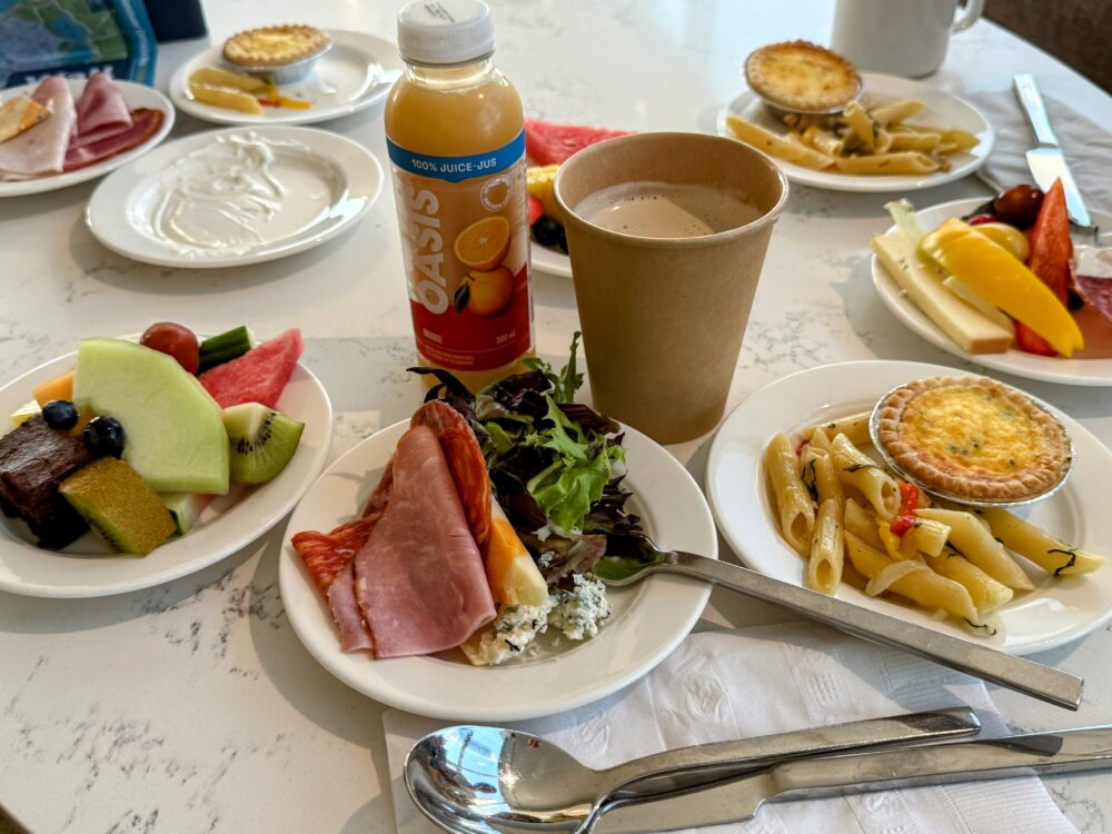 ラウンジでの軽食の盛り付け例。ペンネパスタ、ミニキッシュ、ハム、チーズ、サラダ、フルーツが一皿に盛られ、紙コップのコーヒーとボトル入りのオレンジジュース(Oasis)が添えられている。