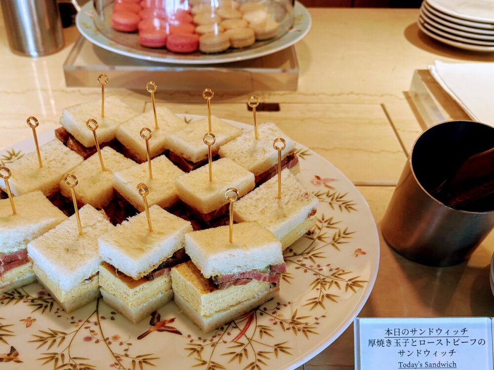 本日のサンドウィッチとして提供された厚焼き玉子とローストビーフのサンドウィッチ。一口サイズにカットされ金のピックが刺さった状態で花柄の皿に整然と並んでいる。奥にはマカロンの皿も見える。