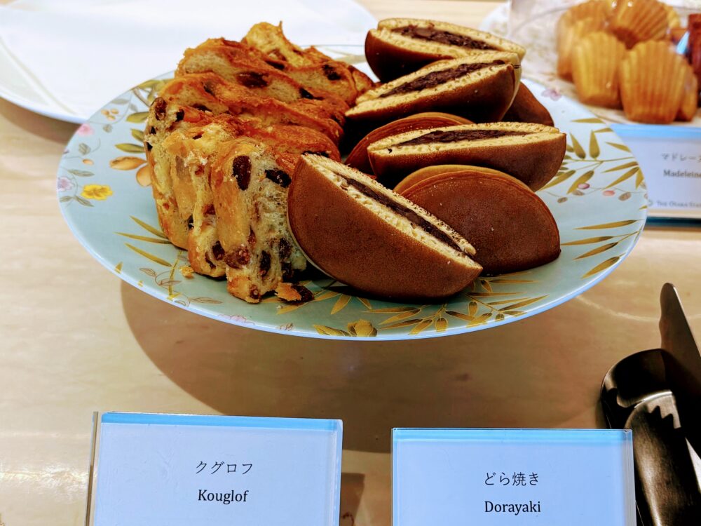 花柄の皿にクグロフとどら焼きが並んでいる。クグロフはレーズン入りで断面が見えるようにカットされており、どら焼きはあんこが見える状態でカットされている。手前にメニュー名のプレートが置かれている。