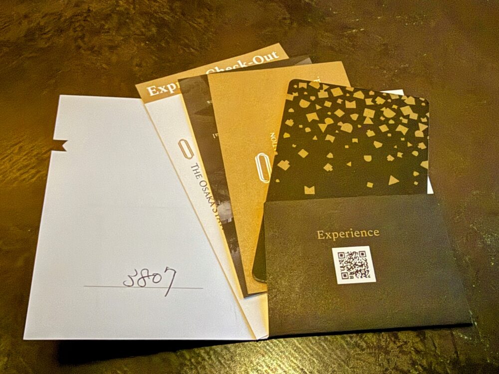 大阪ステーションホテルのチェックイン時に受け取った、部屋番号が書かれた白いキーケース、黒地にゴールドの模様が入ったシックなルームキー、およびエクスプレスチェックアウトや館内案内のカード類。