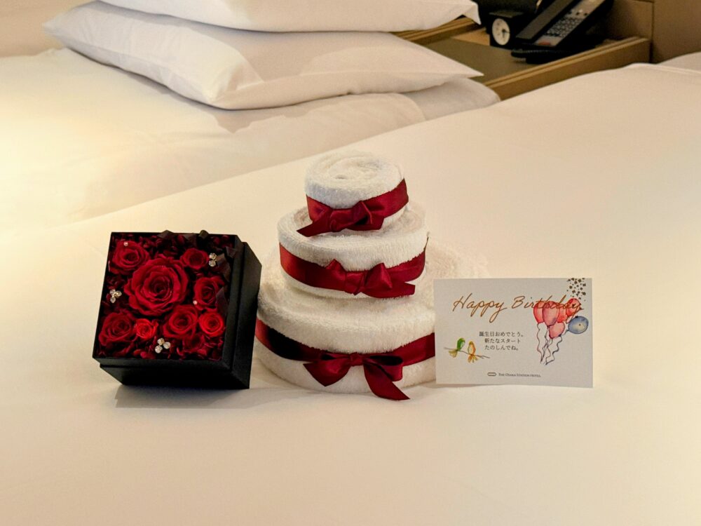 ベッドの上に置かれたお誕生日サプライズのギフト。赤いリボンが巻かれた3段のタオルケーキ、赤いバラのフラワーボックス、メッセージカード。
