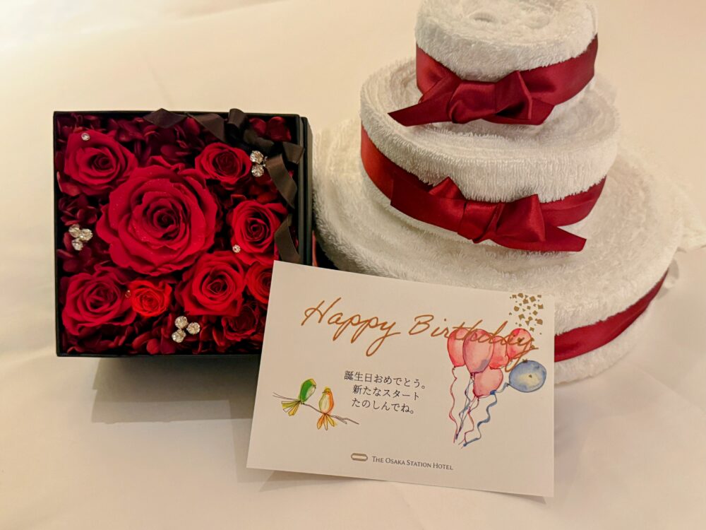 誕生日ギフトのアップ写真。真っ赤なプリザーブドフラワーのボックス、タオルケーキ、「Happy Birthday」と書かれた小鳥と風船のイラスト付きカード。