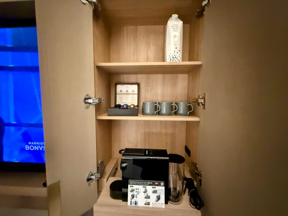 木目調のキャビネット。上段に大きな紙パックの水、中段にネスプレッソのカプセルとマグカップ、下段にコーヒーメーカーが設置されている。