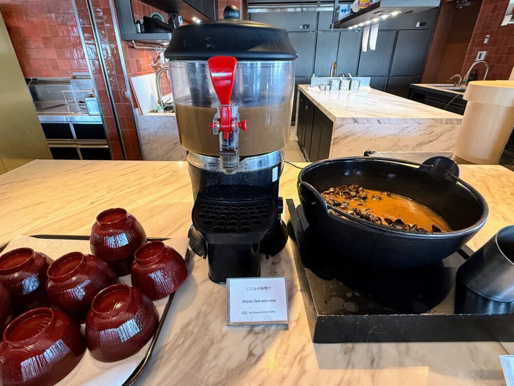 黒い専用ディスペンサーに入った「しじみの味噌汁」と、手前に並べられた赤いお椀。隣の黒い鉄鍋にはしじみがたっぷり用意されている。