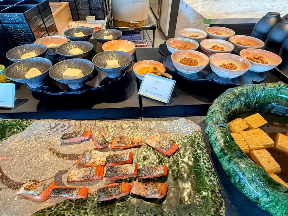 和食ビュッフェ台の様子。手前にこんがり焼かれた鮭、奥には小鉢に入った冷奴や納豆、右側には厚揚げの煮物などが並んでいる。