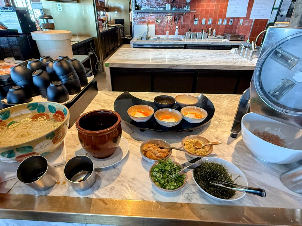 麺類のコーナー。つゆや温泉卵のほか、ネギや刻み海苔などの薬味が並び、右手前には本格的なかつお節削り機が設置されている。