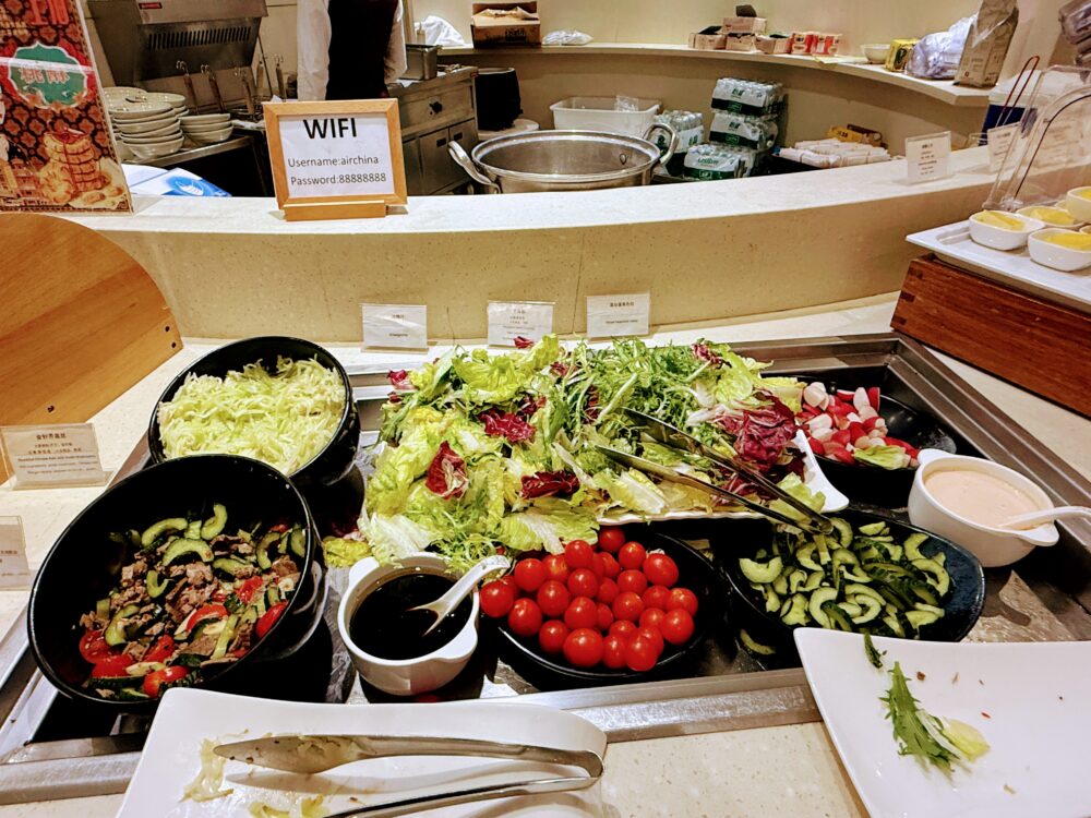 No.71ラウンジのサラダコーナー。レタス・ミニトマト・きゅうりなど新鮮野菜が並ぶ。カウンターにWiFiのユーザー名とパスワードが記載された案内カードが置かれている。