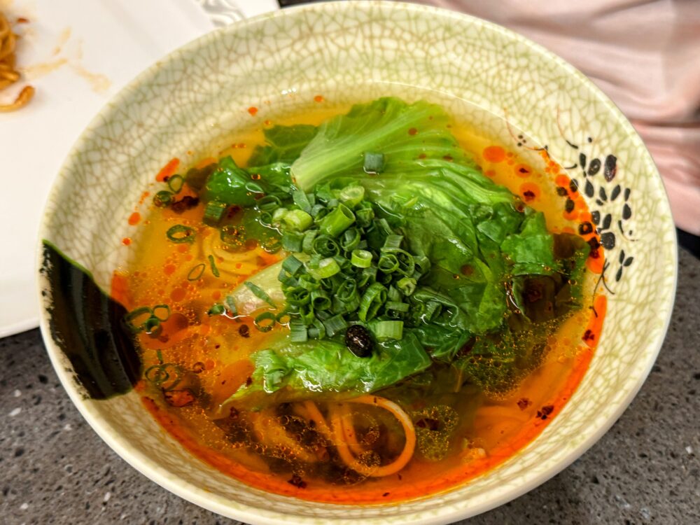 No.71ラウンジLIVEキッチンの麺料理。ひび割れ模様の丼に麺と青菜が入り、赤い辛味スープにたっぷりのねぎと香菜がのっている。