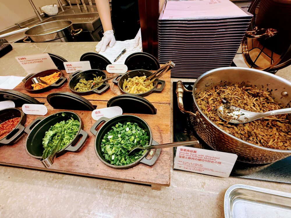 No.72ラウンジLIVEキッチンの薬味カウンター。葱花・香菜・搾菜・酸豆角・紅油竹の子などのトッピングが小鍋に並び、奥で調理スタッフが作業中。「雪菜肉丝麺」のメニュー説明が日本語・英語・中国語で表示されている。