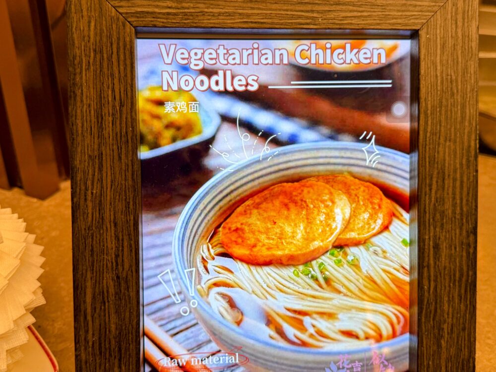 No.72ラウンジLIVEキッチンのメニュー写真。「Vegetarian Chicken Noodles(素鶏麺)」の完成イメージ。揚げ豆腐がのった麺料理の写真が額縁に入って展示されている。