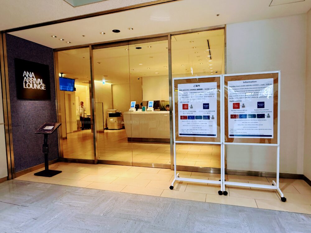 成田空港第1ターミナル南ウイング1階にあるANA ARRIVAL LOUNGEの入口。左側に黒いサインボード、正面にガラス張りの自動ドア、右側に日本語と英語で記載された利用案内のスタンドボードが設置されている。