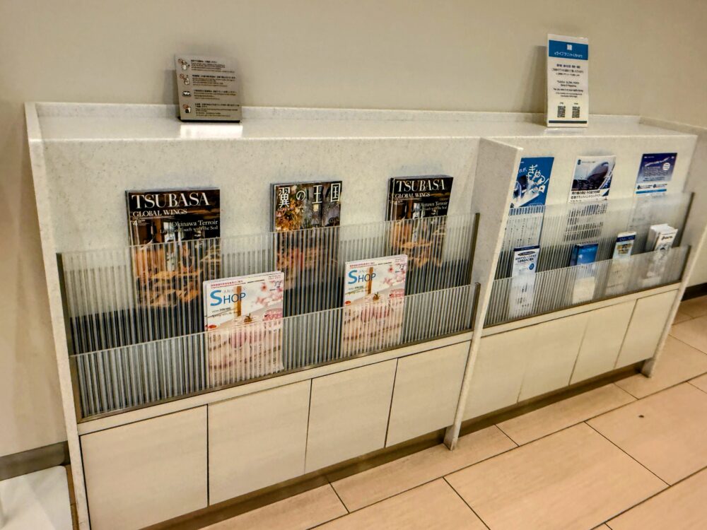 ラウンジ内の雑誌・パンフレットラック。左側にはANA機内誌「TSUBASA GLOBAL WINGS」と「翼の王国」、ANAショッピングカタログが並んでいる。右側にはANA関連のパンフレット類が収納されている。上部にはWi-Fiパスワードの案内カードとQRコード付きのサービス案内が置かれている。