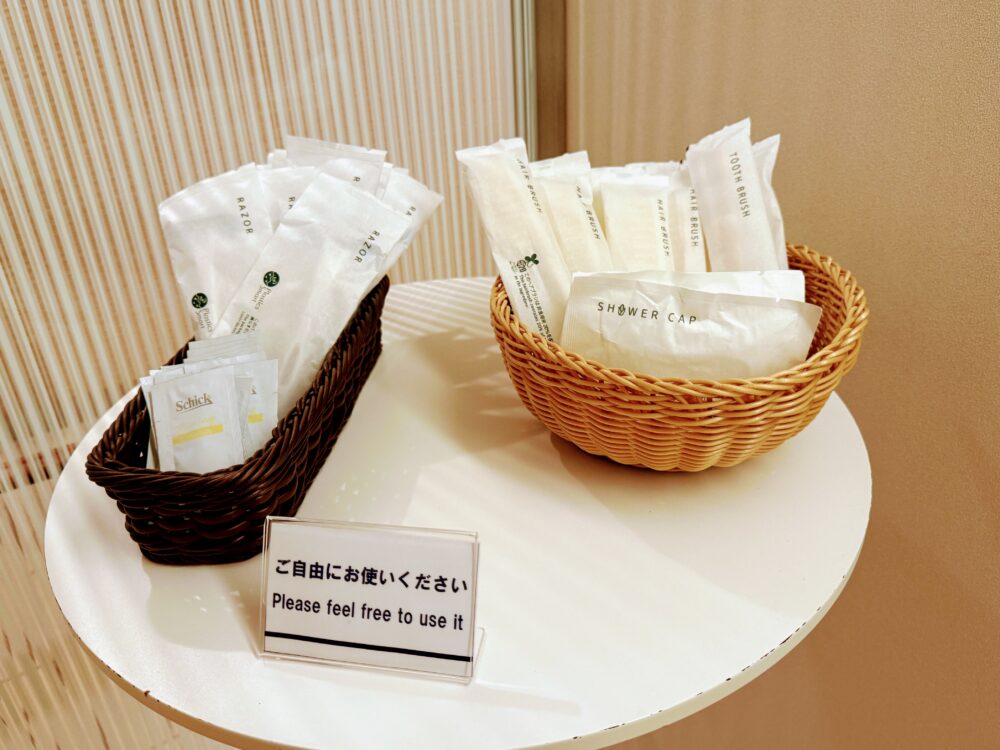 共用エリアのアメニティテーブル。白い丸テーブルの上に、Schickのカミソリやシェービングジェルが入ったダークブラウンのかごと、歯ブラシ・ヘアブラシ・シャワーキャップが入ったナチュラルカラーのかごが並んでいる。手前に「ご自由にお使いください」と書かれた案内カードが置かれている。