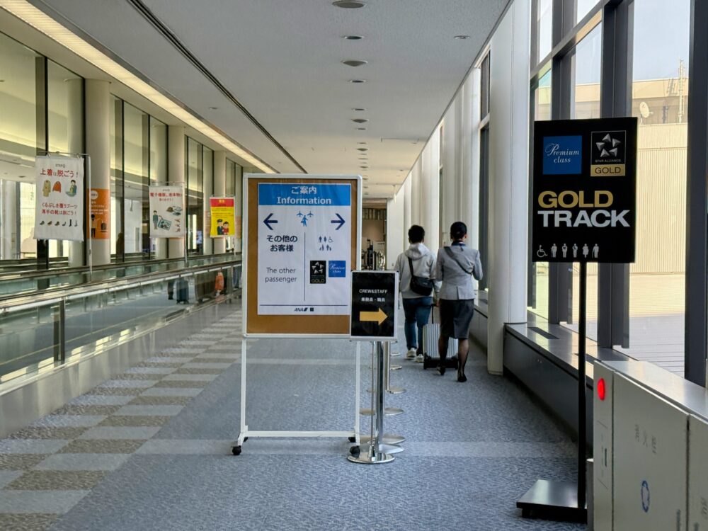 成田空港国内線の保安検査場入口エリア。中央に「ご案内 / Information」と書かれたスタンドボードがあり、一般旅客は左方向へ、プレミアムクラス・Star Alliance GOLDメンバーは正面へ、乗員・職員は右方向へと案内している。右側には黒い「GOLD TRACK」のサインボードが設置されており、プレミアムクラスおよびStar Alliance GOLD会員向けの優先保安検査レーンであることが示されている。奥にスーツケースを引く旅客の姿が見える。