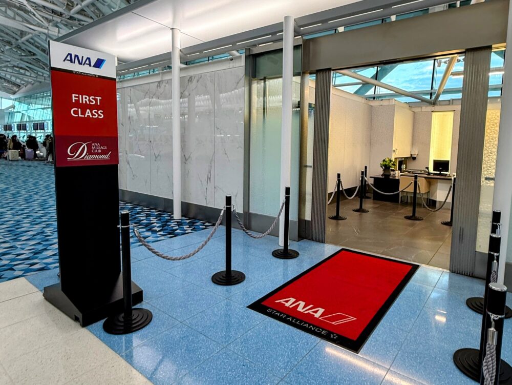 羽田空港のANA SUITE CHECK-IN入口。「FIRST CLASS / ANA Mileage
Club Diamond」と記された赤黒のサインポールと、ANA・Star Allianceロゴ入りの赤いレッドカーペットが入口に敷かれ、ロープパーティションの
奥にチェックインカウンターが見える。