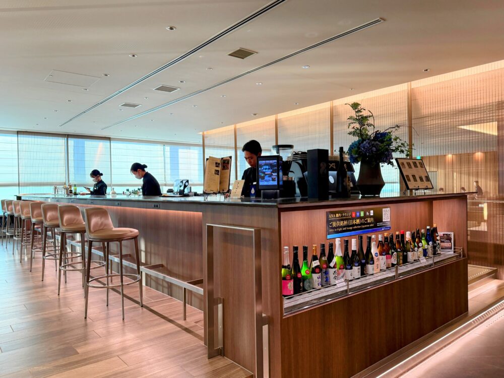 ANA SUITE LOUNGEのバーカウンター。木目調のカウンターにハイチェアが並び、右手には日本酒の小瓶が多数ディスプレイされている。
スタッフ3名が作業中で、奥には空港の景色が見える大きな窓が広がる。