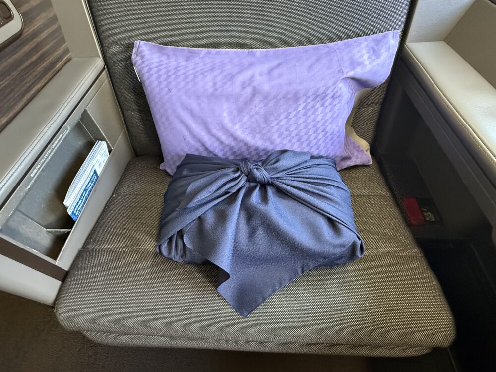 ANA THE Suite のシートに置かれた風呂敷包み。
紺色の風呂敷が上品な結び目でまとめられ、
背後にはラベンダー色のピローが見える。