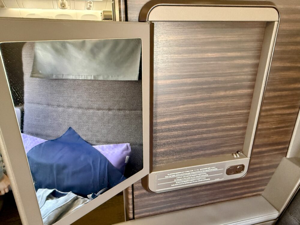 ANA THE Suite 1A席の個室ドア内側。半開き状態のドアに
鏡が取り付けられており、鏡越しに風呂敷に包まれた
パジャマとラベンダー色のピローが映り込んでいる。
木目調のドアパネルには離着陸時の注意書きが記されている。