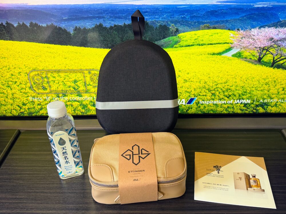 ANA THE Suite のアメニティ一式。43インチモニターを背景に、
キャメル色のETTINGER LONDONコラボポーチ、天然名水のボトル、
黒いSONYヘッドホンケース、サントリー響100周年ウイスキーの
案内カードがサイドテーブルに並んでいる。