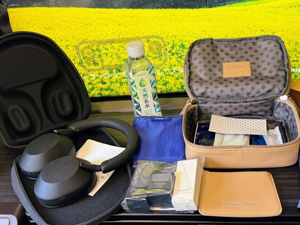 ETTINGER × ANA コラボアメニティポーチを開いた状態。
ポーチ内にはSENSAIのローション・クリーム・リップトリートメントが
収められ、隣にSONYのノイズキャンセリングヘッドホン、
ANAロゴ入りの紺色ポーチが並んでいる。