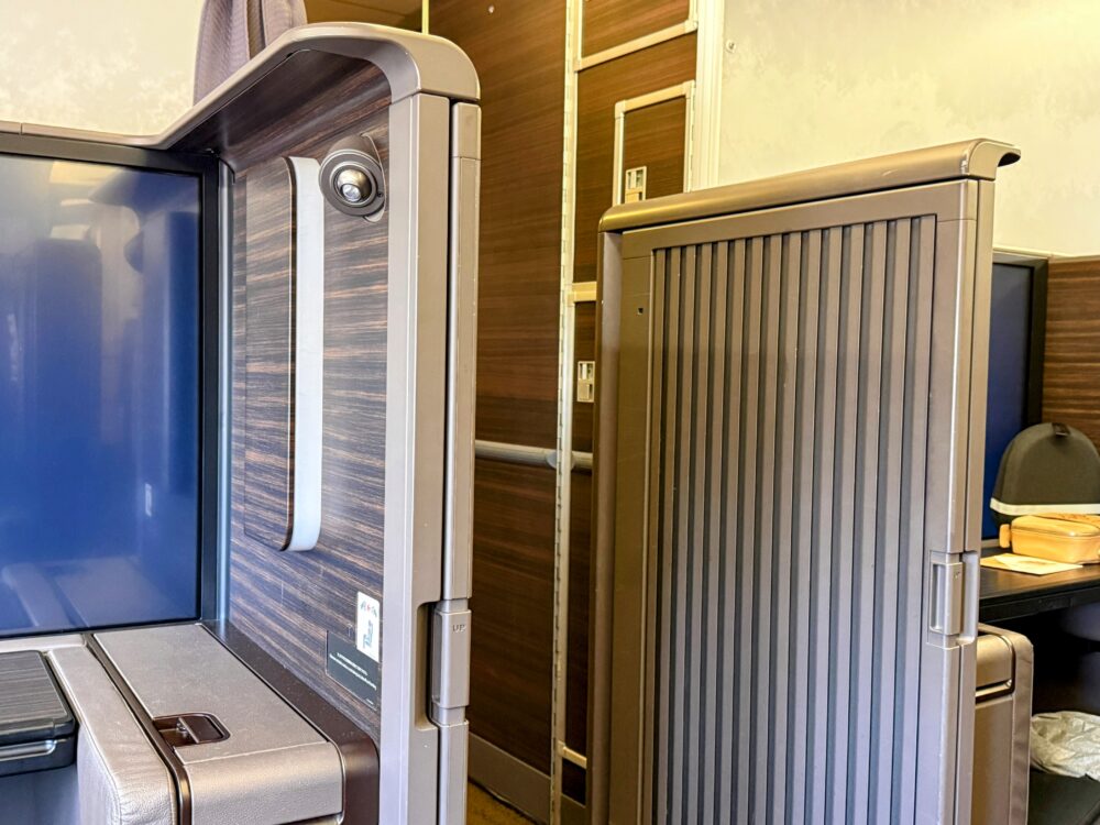 ANA THE Suite 1A席の個室スライドドアが開いた状態。
木目調のパネルとグレーのドアが並び、
奥にはCAが通る通路と隣席のスペースが見える。
右奥にはETTINGERのアメニティポーチが確認できる。