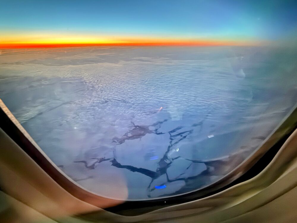 機窓から見た北極圏上空の風景。眼下には割れた流氷が広がり、水平線には鮮やかなオレンジとブルーのグラデーションで日が沈んでいく。
眼下に広がる流氷と、水平線に沈む夕日。この景色だけでも、窓際席を諦めなくてよかったと思いました。
