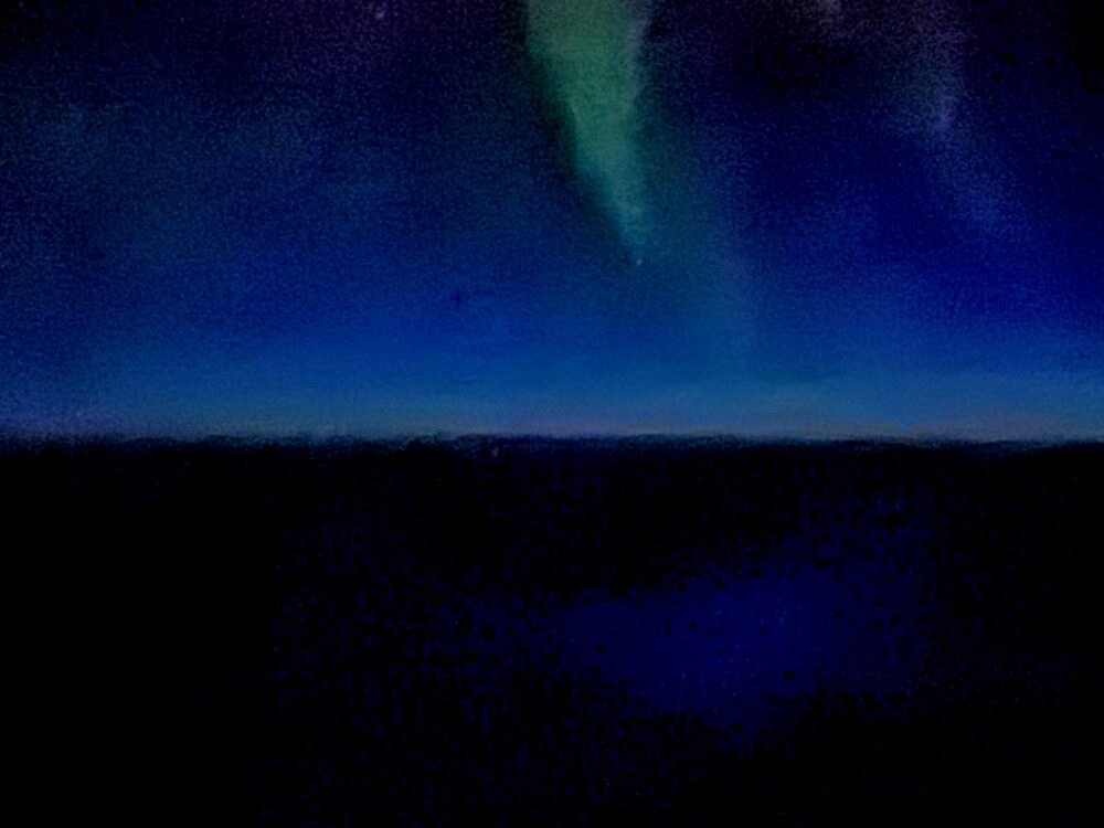北極圏上空の機窓から撮影したオーロラ(1枚目)。深い紺色の夜空に、緑色のオーロラの柱が天頂に向かって縦に伸びている。地平線付近はうっすらと明るい。
iPhoneのカメラを通すと、はっきりと緑に見えたオーロラ。肉眼では白く輝いていました。