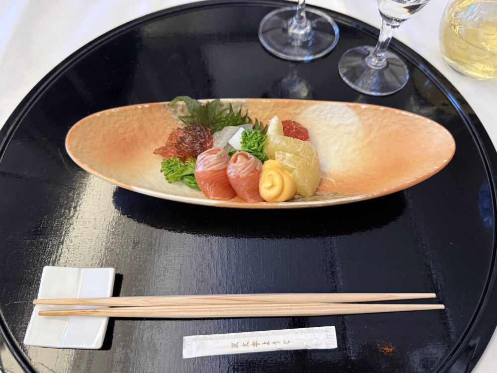 オレンジ色のグラデーションが美しい細長い器に
盛られた旬の刺身盛り合わせ。まぐろ、白身魚、
いくらなどが丁寧に盛り付けられ、黒い丸盆の上に置かれている。
