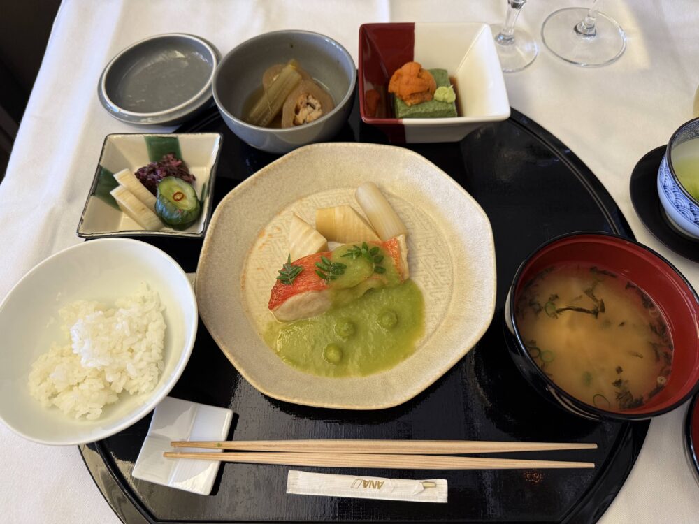 黒い丸盆に盛られた和食コース一式。中央の白い器に
金目鯛の油焼きとウドの炊き合わせ、青豆ソース添え。
左に小鉢と漬物、右奥に前菜の小皿、右手前に赤い椀の味噌汁、
左手前に白飯が並ぶ。