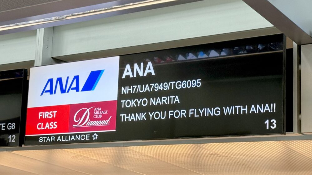 空港の搭乗口付近に設置されたANAのフライト情報ディスプレイ。「ANA NH7/UA7949/TG6095 TOKYO NARITA THANK YOU FOR FLYING WITH ANA!! 13」と表示されており、成田空港行きのフライトであることが示されている。左側にはANAロゴ、ファーストクラスの赤いバッジ、ANAマイレージクラブ・ダイヤモンドのロゴ、スターアライアンスのロゴが表示されている。