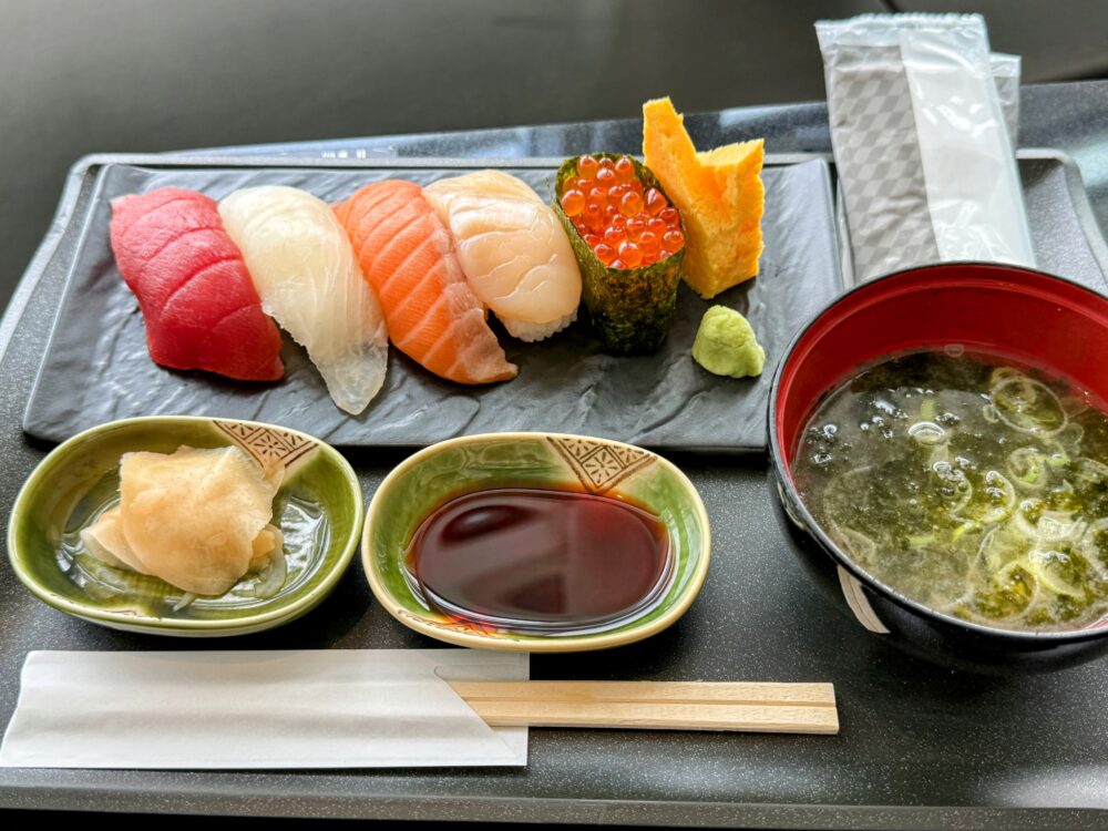 ANAスイートラウンジSUITE DININGのお寿司。まぐろ、白身、サーモン、ほたて、いくら軍艦、玉子の握り寿司がスレート皿に並び、わさび・醤油・がり・わかめ汁添え（第3ターミナルにて撮影）。
