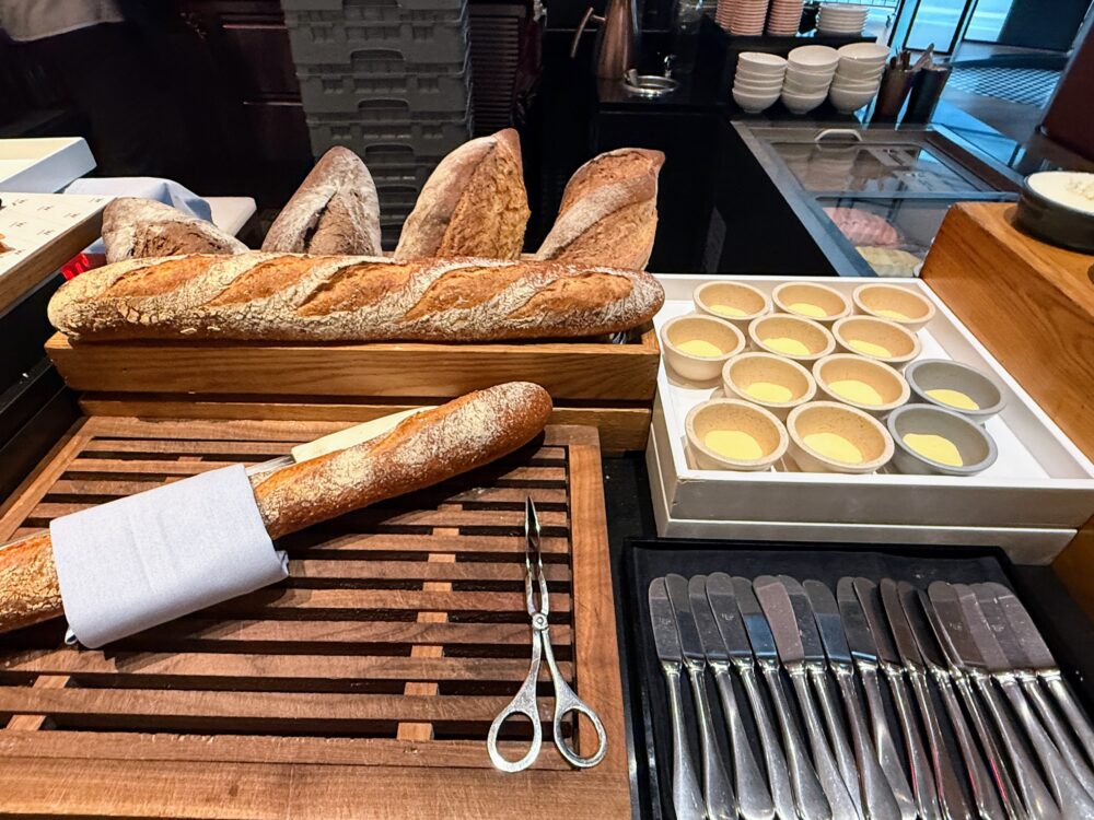 W上海「THE KITCHEN TABLE」朝食ビュッフェのパンコーナー。バゲットとフランスパンがウッドトレイに並び、横には卵タルト(エッグタルト)の小皿も置かれている。