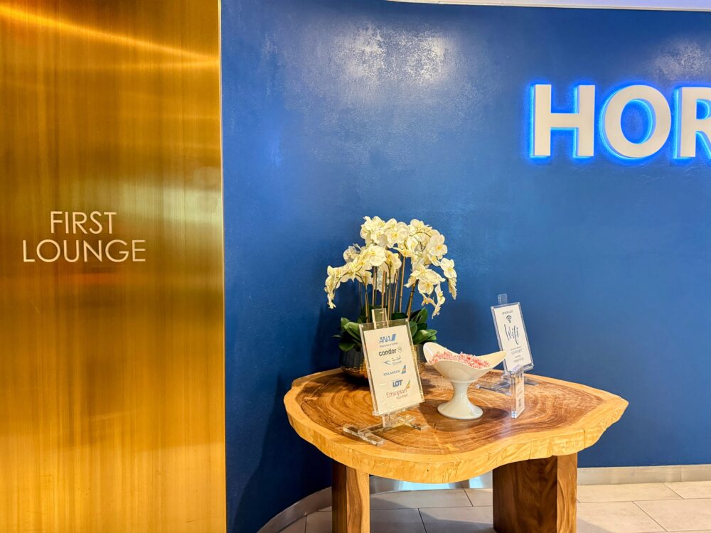 ファーストクラスラウンジの入口サイン「FIRST LOUNGE」。ゴールドのプレートと隣のHORIZONS T7 LOUNGEのロゴウォール、木製テーブルのウェルカムディスプレイが見えます。