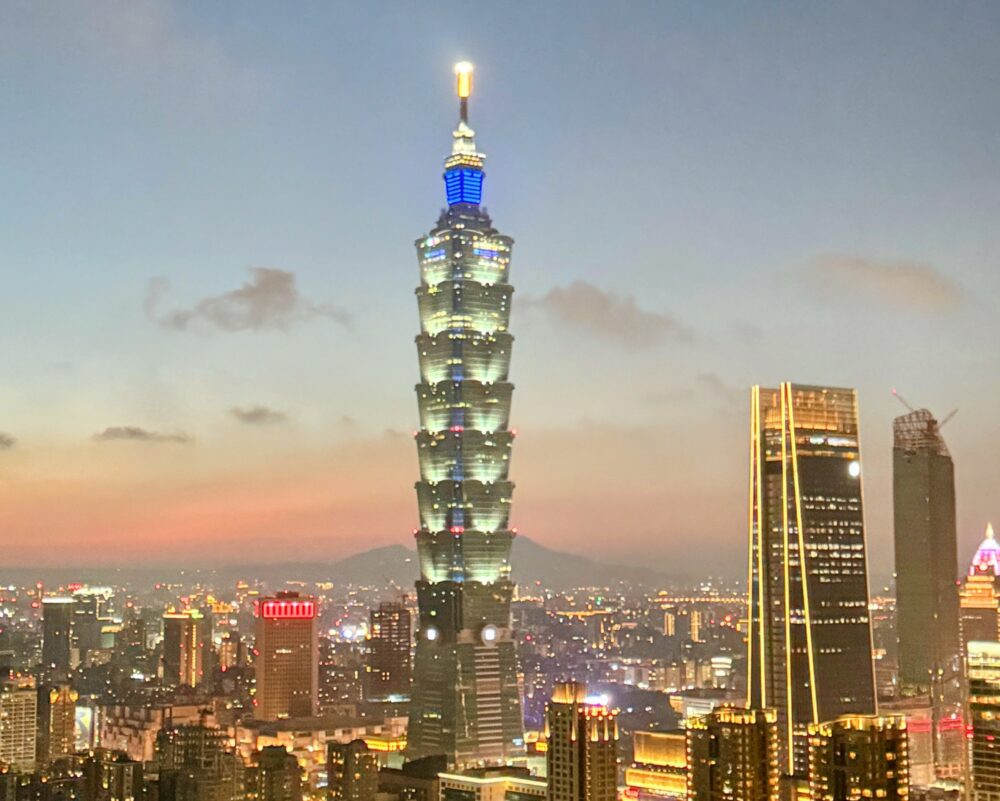 夕暮れ時の台湾・台北の街並み。ライトアップされた象徴的な超高層ビル「台北101」を中心に、周囲のビル群の明かりが輝き、幻想的な雰囲気が漂う都市の夜景。