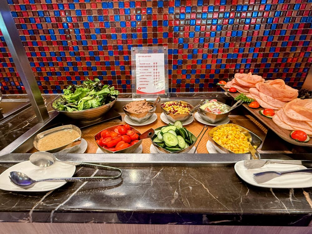 SATSプレミアラウンジの新鮮な野菜やハムを自由にトッピングできるDIYサラダバーと、横に並ぶエビせんべい。