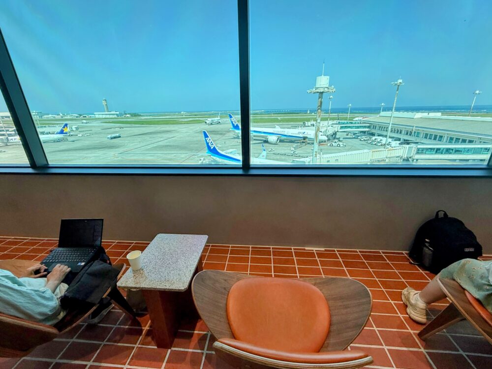那覇空港カードラウンジ「LOUNGE HANA」の大窓から滑走路とANA機を見渡せるオレンジ色のラウンジ席でノートパソコン作業をする様子