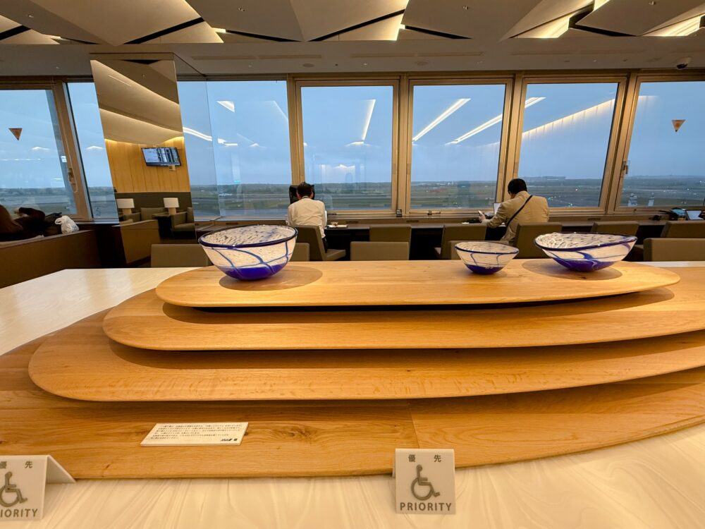ミズナラ材の大テーブルに、北海道の空と海をイメージした青い小樽のガラス工芸が飾られたANAスイートラウンジの席。窓の外には滑走路が広がる。