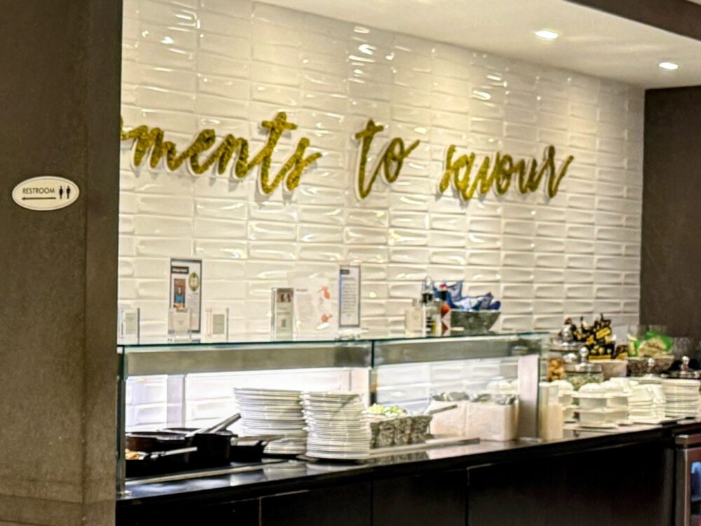 ビジネスクラスラウンジのフードカウンター入口。「moments to savour」のゴールドの文字が白いタイル壁に飾られ、カウンター上には白いプレートが積まれています。