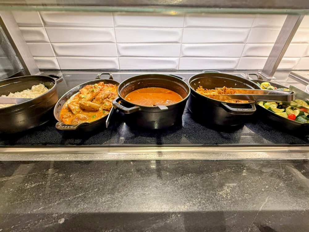 ビジネスクラスラウンジのホットミールコーナー。チキンカレー、トマトスープ、チキンライス、ズッキーニソテーなどが黒い鋳鉄鍋に入って温められています。