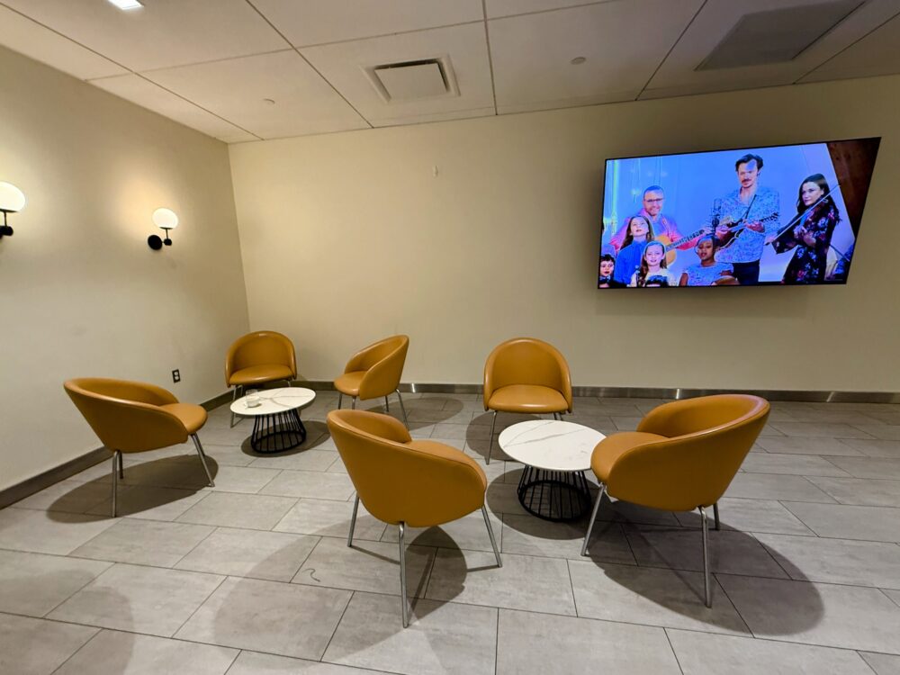 ビジネスクラスラウンジのテレビルーム。オレンジのラウンジチェアが半円形に並び、正面には大型テレビが設置されています。