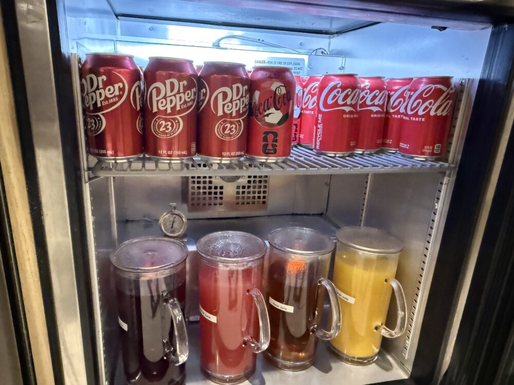 ファーストクラスラウンジの冷蔵庫内。上段にDr PepperとCoca-Colaの缶、下段にはグレープ・クランベリー・オレンジなどのフルーツジュースが入った透明ピッチャーが冷やされています。