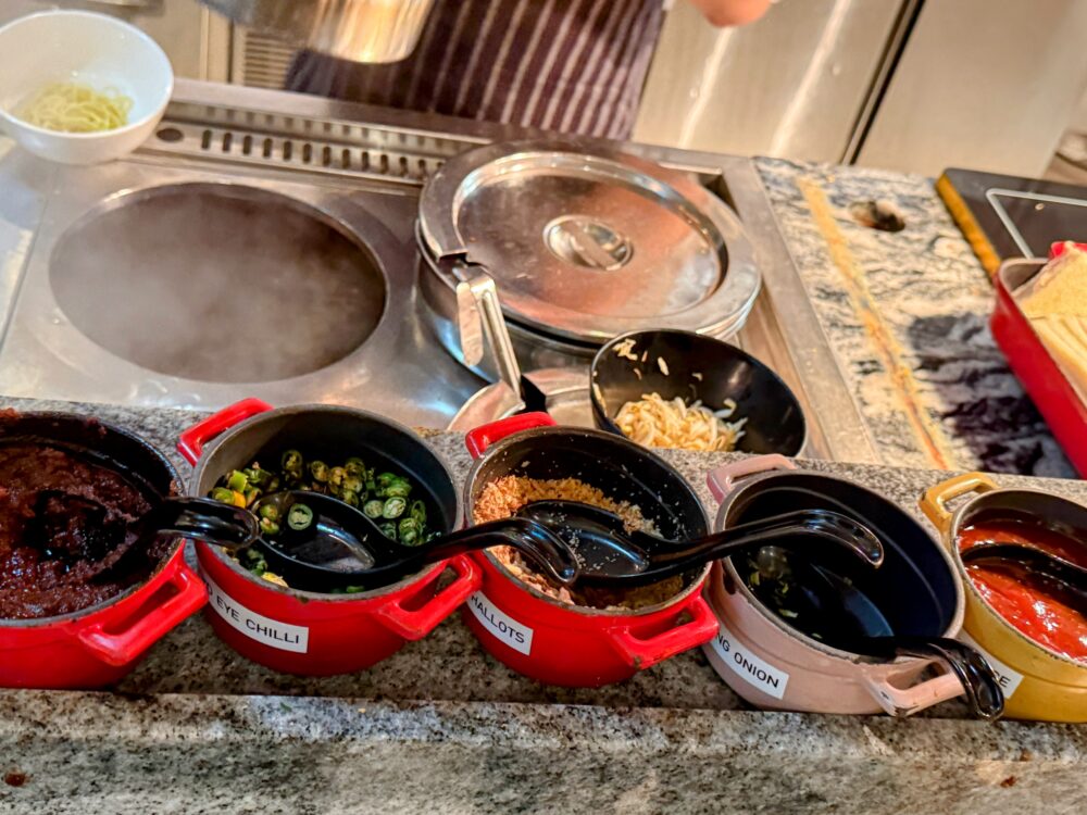 ライブキッチンカウンターに並ぶ、チリ、フライドオニオン、揚げ玉などの麺料理用トッピング