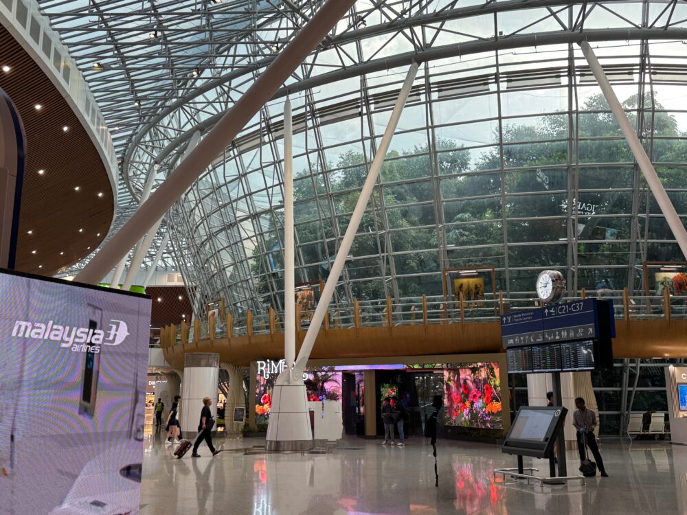クアラルンプール国際空港（KLIA）サテライトターミナル中央の吹き抜けエリア。左手にマレーシア航空の大きなデジタルサイネージ、中央奥に屋内庭園「RIMBA（ジャングルボードウォーク）」の入口、右手にはフライト情報ディスプレイと案内板が設置されている。