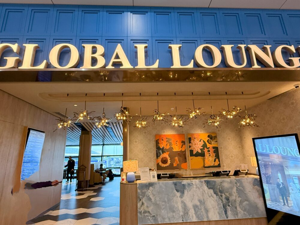 「GLOBAL LOUNGE」のエントランス。鮮やかな青い壁面にゴールドの立体的な文字看板が掲げられ、華やかなペンダントライトと受付カウンター、美しい絵画が配置されている。