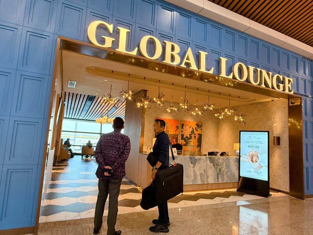 「GLOBAL LOUNGE」の受付カウンター前。バティック柄のシャツを着た男性客がスタッフと話しており、隣には大きなバッグが置かれている。右手のモニターにはスパの案内が表示されている。
