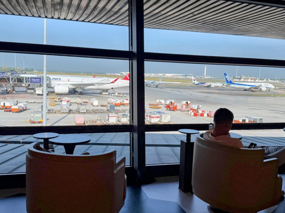 「GLOBAL LOUNGE」の窓際席から見える滑走路の景色。手前のラウンジ内には、革張りのアームチェアに座ってスマートフォンを見る乗客の姿があり、大きな窓の外には駐機中の航空機やターミナル、エプロンエリアが広がっている。