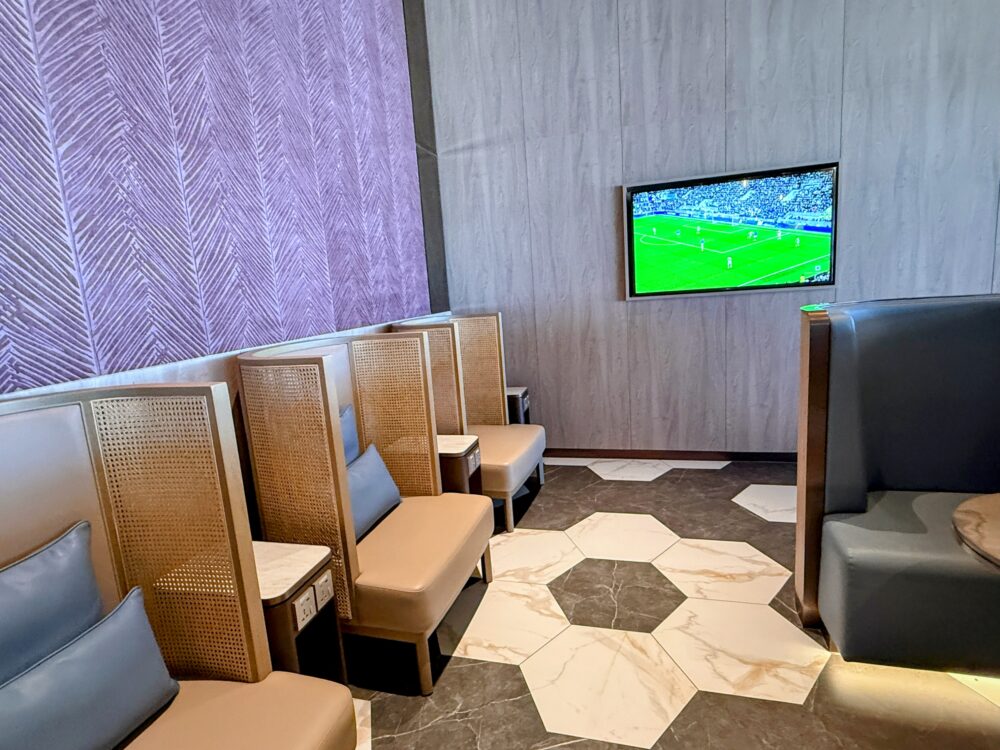 「GLOBAL LOUNGE」のTVエリア。紫色の植物柄の壁面とグレーの壁に囲まれた空間に、青いクッション付きのソファ席が配置されており、壁に掛けられたモニターにはサッカーの試合映像が映し出されている。