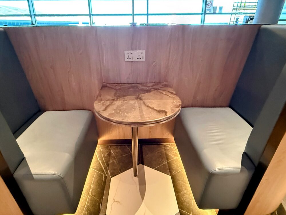 「GLOBAL LOUNGE」内のボックス型ソファ席。木目のパネルで囲まれた対面式のグレーのソファ席で、中央には大理石調の丸テーブルが設置されている。背面の上部には電源コンセントが備えられている。
