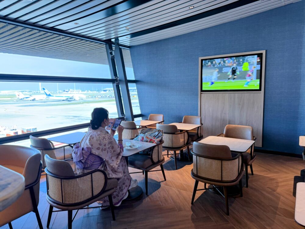 「GLOBAL LOUNGE」の窓際に面したダイニングエリア。青い壁と木目の天井が特徴的な空間で、着物姿の女性が席に座りスマートフォンを操作している。窓の外には駐機中の航空機が見え、壁のモニターではスポーツ中継が流れている。
